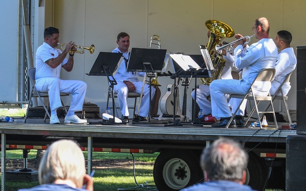 A small brass ensemble of four musicians dressed in white naval uniforms performs on an outdoor mobile stage. Two audience members, seen from behind, watch the performance while seated at a red picnic table on a grassy lawn. The setting appears to be an outdoor public event, likely a military band concert.