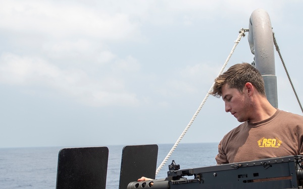 A young male sailor in a brown 'RSO' t-shirt and blue trousers is working on a mounted crew-served weapon system on the deck of a naval vessel at sea. He appears to be performing maintenance or inspection on what appears to be an M2 .50 caliber heavy machine gun mounted on a shipboard gun mount with blast shields. The ocean and overcast sky are visible in the background.