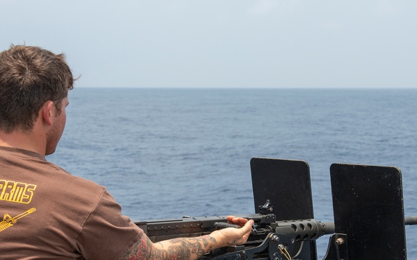 A male individual wearing a brown 'Small Arms Instructor' t-shirt is positioned at a mounted machine gun on the deck of a naval vessel. He is facing outward toward open ocean, appearing to man or inspect the weapon system. The setting is a clear day at sea with calm blue water visible in the background.