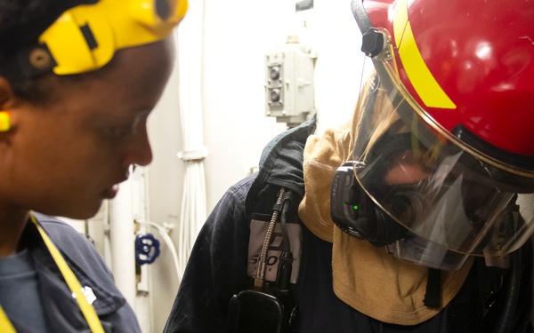 Two personnel, likely military or naval crew members, are reviewing a document or checklist together in what appears to be a ship's interior compartment. One person is wearing full firefighting or damage control gear including a red helmet with yellow markings, a full-face respirator mask, and white gloves, while writing on a form. The other person is in standard duty uniform with yellow safety accessories including ear protection and a lanyard.