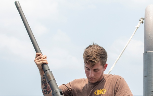 A young male sailor in a brown 'RSO' t-shirt and navy blue work pants is performing maintenance or assembly on a crew-served weapon, likely an M2 .50 caliber heavy machine gun, mounted on the deck of a naval vessel. The setting is an open ocean environment with calm seas visible in the background. The individual appears focused on the task, handling the barrel of the weapon.