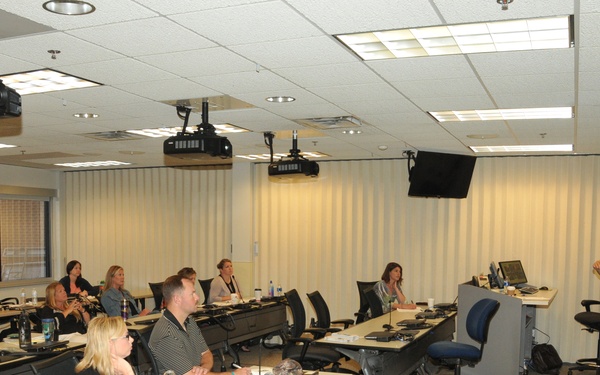 A professional training or educational session is taking place in a modern conference/classroom setting. A male presenter stands at the front near a projection screen, addressing approximately 15-20 seated attendees arranged at tiered desks. The room is well-equipped with multiple ceiling-mounted projectors, a wall-mounted monitor, and a podium.