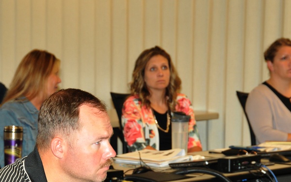 A group of adults are seated at tables in what appears to be a meeting room or conference setting. A man in a black and white striped polo shirt is prominently in the foreground, with four women visible in the background. All individuals appear to be attentively focused on something outside the frame, likely a presentation or speaker.