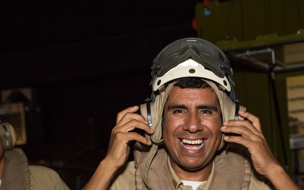 Two military personnel are photographed in what appears to be a naval vessel interior. The foreground subject is smiling broadly while adjusting a flight deck helmet with integrated hearing protection. Both individuals are wearing tan/khaki uniforms with flotation vests and flight deck helmets.