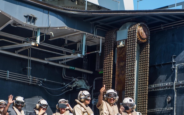 A group of personnel wearing flight deck safety gear — including white helmets, goggles, hearing protection, and float coats — are gathered on what appears to be the deck or edge of a large naval vessel. The individuals are dressed in a mix of tan flight deck uniforms, civilian clothing, and military camouflage, suggesting a mix of military personnel and possibly civilian visitors or media. One person in the center gives a thumbs-up gesture while others observe or photograph an off-camera event.