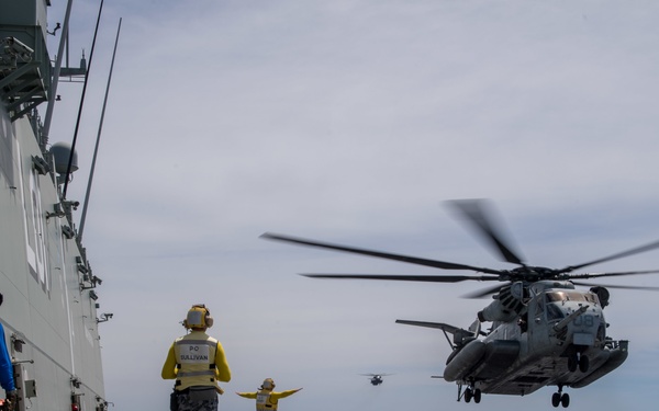 A large military helicopter, appearing to be a CH-53E Super Stallion, is landing or hovering over the flight deck of a naval vessel. Deck crew members in colored safety vests are directing the aircraft, with at least one additional helicopter visible in the background. The scene appears to be a routine flight deck operations exercise, likely involving multinational naval forces based on the visible insignia and vest markings.