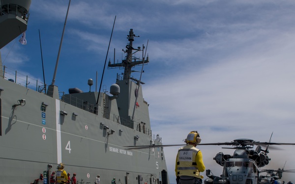 A military helicopter operation is underway on the flight deck of a naval vessel, with the hull number 'L01' prominently visible on the ship's superstructure. Personnel in high-visibility yellow vests and military uniforms are managing flight deck operations, with at least one helicopter visible with rotors spinning. The scene appears to be an Australian Navy vessel operating at sea under partly cloudy skies.