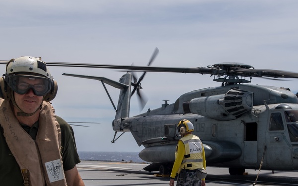 The image shows a flight deck operation aboard what appears to be a naval vessel, with a large military helicopter (CH-53E Super Stallion) on the deck with rotors spinning. Personnel in flight deck safety gear, including helmets, goggles, and life vests, are present on the deck. A deck handler in a yellow vest and camouflage pants is observed near the helicopter.