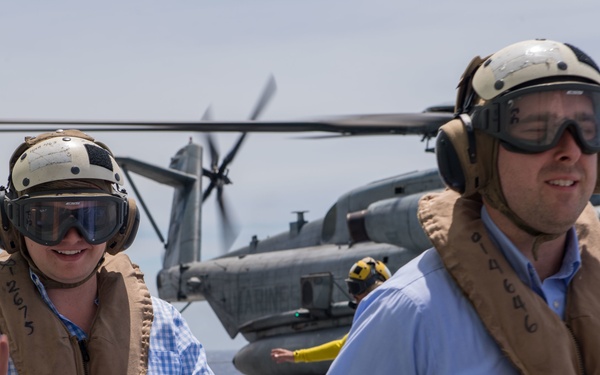 Several civilians are walking on what appears to be a naval vessel flight deck, wearing safety equipment including helmets, goggles, and inflatable life vests. A military tiltrotor aircraft, identifiable as an MV-22 Osprey, is visible in the background with its rotors visible. The scene suggests a civilian tour or official visit to a military ship.