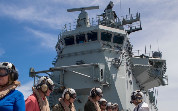 A group of visitors and military personnel are standing on the flight deck of a large naval vessel, likely an amphibious assault ship, wearing safety helmets and life vests. A person in a white shirt and camouflage trousers — possibly an Australian Navy crew member — is greeting or welcoming the group with handshakes. Helicopters are visible in the background on the flight deck, and the ship's island superstructure dominates the background.