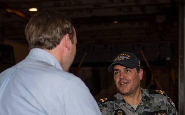 A military officer in Australian Navy camouflage uniform is shaking hands with a civilian in a light blue shirt, likely aboard a naval vessel. The officer's name tag reads 'ISOLA ELIAS' and his cap bears markings consistent with HMAS Adelaide (LHD 01). The setting appears to be an interior deck or hangar bay of a warship.