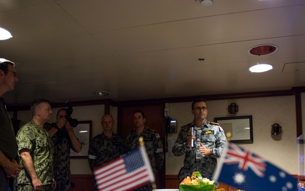 A formal military reception or dining event is taking place in what appears to be a ship's wardroom or mess hall. Both United States Navy and Royal Australian Navy personnel are present, along with civilians, gathered around a buffet table decorated with American and Australian flags. A senior Australian naval officer appears to be addressing the group.