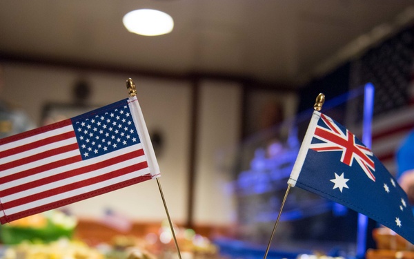 The image shows a formal reception or diplomatic event featuring both American and Australian flags displayed on a table alongside miniature ship models labeled 'HMAS Adelaide' and 'USS Adelaide'. The table is set with various food items suggesting a buffet-style gathering, likely a military or naval bilateral event. The presence of both nations' flags and matching ship models indicates a joint US-Australian naval ceremony or commemorative occasion.