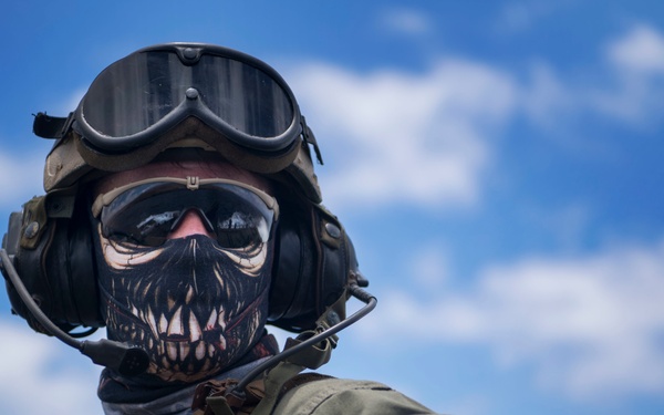A military personnel member is photographed from a low angle while positioned in or on a military vehicle. The individual is wearing tactical gear including a combat helmet with mounted goggles, communications headset, tactical vest, and a skull-patterned face covering/gaiter. The image is taken against a bright blue sky with scattered clouds.