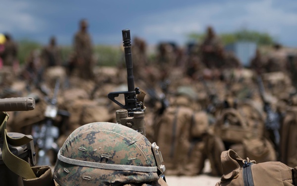 The image shows a large assembly of military personnel with their gear staged on sandy ground. In the foreground, multiple tactical backpacks, rifles, and a MARPAT (Marine pattern) camouflage helmet are clearly visible. The background shows numerous soldiers and additional equipment in a shallow depth-of-field blur.
