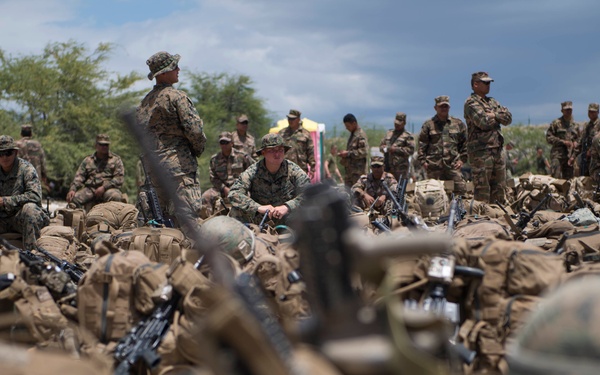 A large group of military personnel in camouflage uniforms are assembled in an outdoor staging area, surrounded by tactical gear, backpacks, helmets, and rifles stacked or laid on the ground. The scene appears to depict a rest or assembly phase during a military exercise or operation. The foreground is intentionally blurred, drawing focus to the personnel gathered in the mid and background.