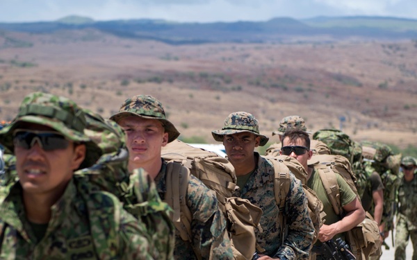 A column of military personnel in full combat gear marches along a dirt road in an arid, hilly landscape. The group appears to include soldiers from at least two different nations, identifiable by distinct camouflage uniform patterns. They are carrying large rucksacks and at least one visible assault rifle, suggesting a field training exercise or patrol movement.