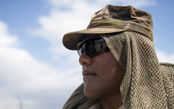 A person wearing military or tactical gear is photographed in an outdoor desert-like environment. They are equipped with a camouflage cap, a mesh/net head covering draped over their shoulders, reflective sunglasses, and a tan/khaki uniform. The image appears to be taken during daylight with a partly cloudy sky in the background.