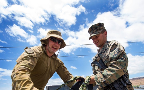 Two military personnel are working together to handle gear and equipment in an outdoor setting near what appears to be a port or coastal facility. One soldier wears a tan/olive uniform with a wide-brim hat and sunglasses, while the other wears MARPAT digital camouflage consistent with US Marine Corps uniform and carries a rifle. A larger group of uniformed personnel is visible in the background.
