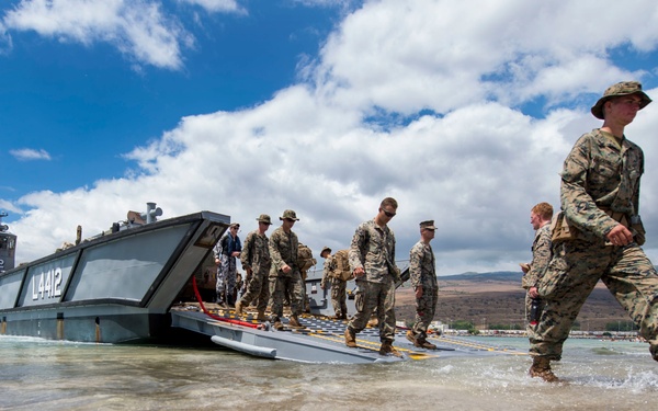 Military personnel in camouflage uniforms are disembarking from a landing craft designated 'L4412' onto a shallow beach. The soldiers are wading through ankle-deep water as they move from the vessel's lowered ramp onto shore. The setting appears to be a tropical or subtropical coastal location with mountains visible in the background.
