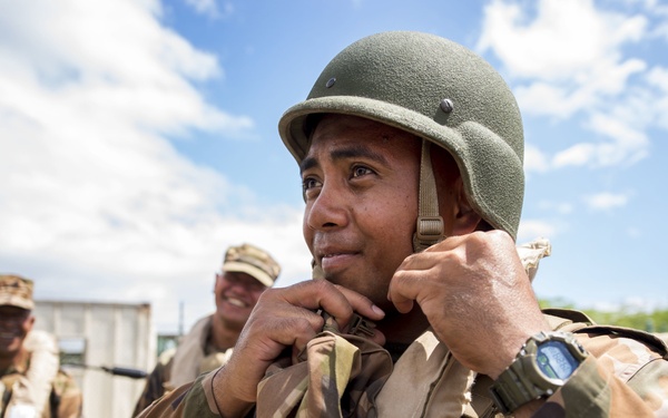 A soldier in the foreground is fastening the chin strap of a green ballistic helmet while wearing camouflage fatigues and a tan tactical vest. Several other military personnel in similar uniforms are visible in the background, some smiling, suggesting a training or preparation scenario. The setting appears to be an outdoor military compound or training area under a partly cloudy sky.