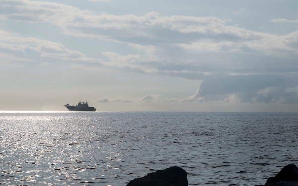 A large naval or military vessel is silhouetted on the horizon of a calm, open sea, photographed from a rocky shoreline or breakwater. The image is backlit by diffused sunlight reflecting off the water's surface, creating a silver-toned, atmospheric seascape. Dark foreground rocks contrast with the luminous water and partly cloudy sky.