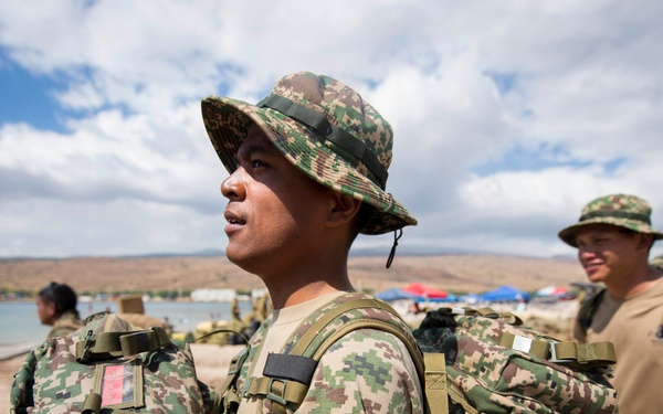 The image shows military personnel in camouflage uniforms and boonie hats carrying large tactical backpacks on a beach setting. At least three soldiers are visible, with the foreground subject wearing a digital camouflage pattern uniform and hat. The setting appears to be a coastal training area with a bay, sandy beach, and tents visible in the background.