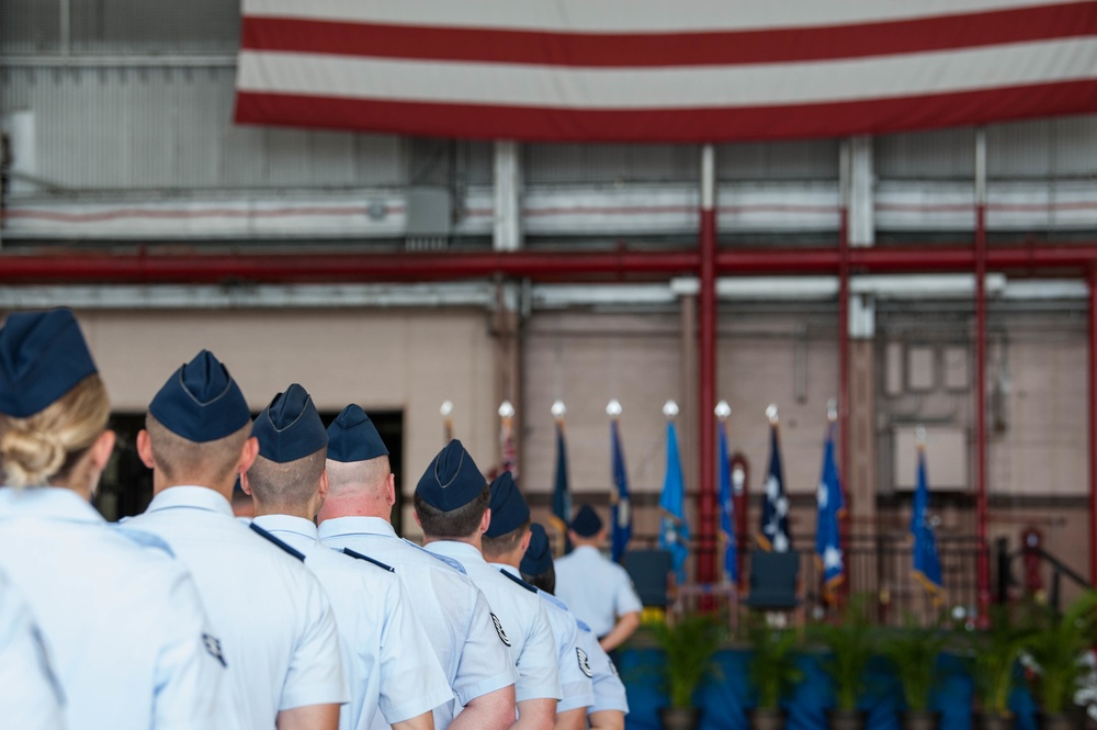A formal United States Air Force military ceremony is taking place inside or in front of a large hangar facility. Personnel in dress blue and white uniforms are standing in formation facing a stage area with multiple flags displayed. A large American flag banner is prominently displayed on the hangar structure in the background.