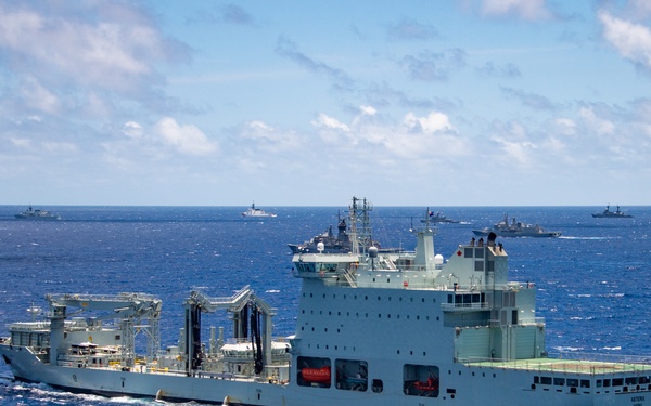 A large naval auxiliary replenishment vessel, painted in light gray/pale green, is underway in open blue ocean waters. Multiple warships are visible in the background, suggesting a multinational naval formation or exercise. The ship appears to be a fleet replenishment oiler or auxiliary support vessel equipped with underway replenishment (UNREP) rigs.