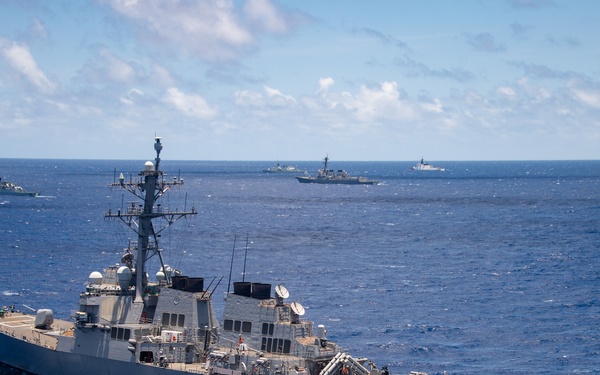 The image shows a U.S. Navy Arleigh Burke-class guided-missile destroyer sailing in open blue ocean waters, likely during a multinational naval exercise or formation transit. Several additional warships from what appear to be multiple nations are visible in the background at varying distances. The weather is clear with partial cloud cover and excellent visibility.