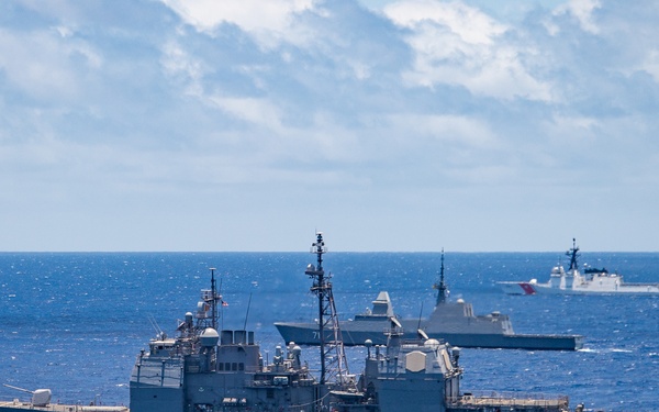 A formation of multiple naval warships is underway on open blue ocean under a partly cloudy sky. The foreground vessel bears hull number 57, identifiable as a US Navy Ticonderoga-class guided-missile cruiser, with additional warships visible in the background including at least one vessel with hull number 71. The scene appears to depict a multinational naval exercise or formation sailing, with ships of varying designs and at least one vessel displaying a red-striped hull consistent with a Coast Guard or foreign navy cutter.
