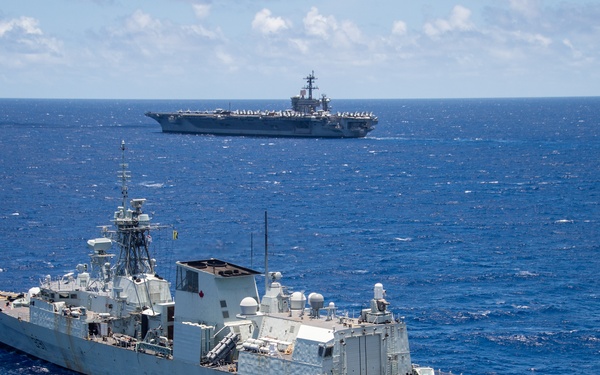 The image shows two naval vessels operating in open blue ocean under partly cloudy skies. In the foreground is a grey frigate bearing hull number 331, and in the background is a large aircraft carrier. The ships appear to be conducting a joint naval exercise or transit operation.