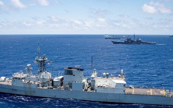 An aerial photograph shows a naval warship bearing hull number 331 underway in open blue ocean waters. A Canadian maple leaf insignia is visible on the superstructure, identifying it as a Royal Canadian Navy vessel. At least two additional warships are visible in the background on the horizon.