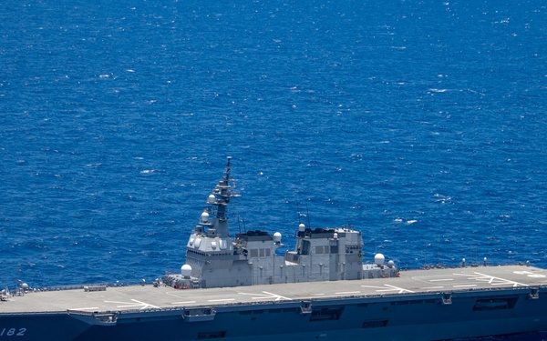 An aerial photograph of a large Japanese Maritime Self-Defense Force helicopter destroyer bearing hull number 182, identified as JS Ise (DDH-182), underway in open blue ocean waters. The vessel features a full-length flat flight deck with multiple helicopter landing spots marked, an island superstructure on the starboard side, and a gray naval paint scheme. The ship appears to be operating independently with no other vessels visible.
