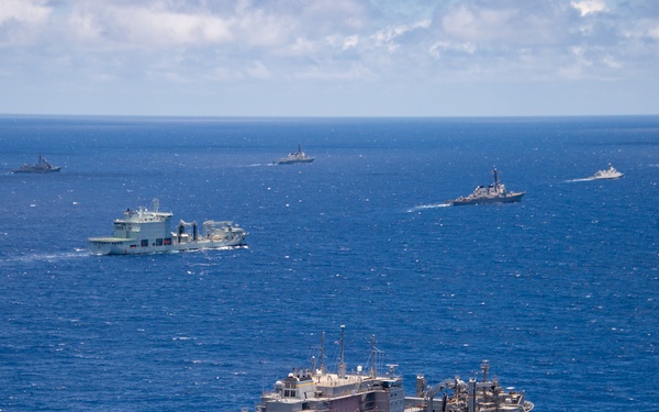 An aerial photograph shows a naval formation of multiple military vessels underway in open blue ocean waters under partly cloudy skies. The foreground features a large gray auxiliary or replenishment ship, with several additional warships and support vessels visible in the middle and background distances. The formation suggests a multinational or fleet exercise at sea.