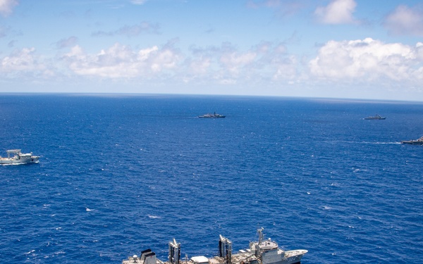 An aerial photograph shows a formation of multiple naval vessels underway in open blue ocean under partly cloudy skies. The foreground features a large replenishment or auxiliary ship with complex superstructure and cranes, while several warships and support vessels are visible at varying distances in the background. The scene appears to depict a multinational or fleet naval exercise or transit formation.