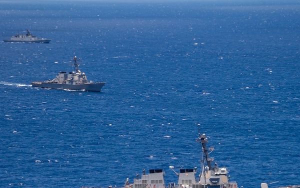 Three naval warships are sailing in formation across open deep-blue ocean waters. The foreground vessel appears to be a U.S. Navy Arleigh Burke-class guided-missile destroyer, with two additional warships visible at increasing distances in the background. The formation suggests a coordinated naval exercise or transit operation.