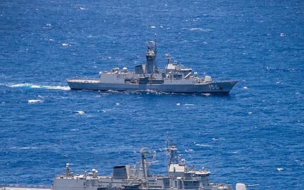 An aerial photograph shows two gray naval warships underway in open blue ocean waters. The background vessel bears the hull number '156' and appears to be a frigate-class warship, while the foreground vessel is a larger warship, possibly a replenishment or amphibious ship. Both vessels are sailing in a parallel formation, suggesting a coordinated naval exercise or transit operation.