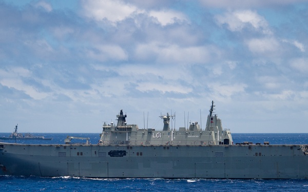 A large amphibious assault ship bearing the hull number 'L01' is underway on open blue ocean waters. The vessel appears to be an Australian Navy Canberra-class Landing Helicopter Dock (LHD), identifiable by its ski-jump flight deck and distinctive hull markings. Additional naval vessels are visible in the distant background, suggesting a multi-ship formation or exercise.