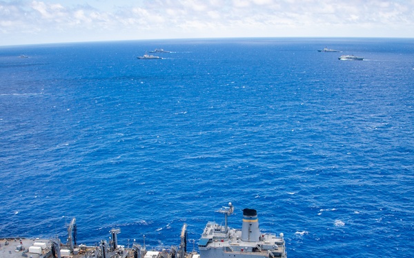 An aerial photograph shows a large naval formation of multiple warships and support vessels operating in open blue ocean waters under partly cloudy skies. The foreground features a prominent gray replenishment or auxiliary support ship with a helicopter landing pad, replenishment rigs, and orange life rafts visible on deck. Several additional naval vessels are dispersed across the background, suggesting a multinational or fleet exercise.