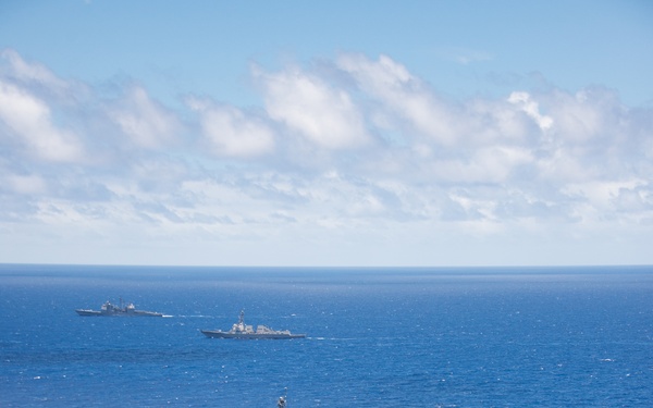 An aerial photograph shows a formation of four naval vessels underway in open blue ocean under partly cloudy skies. The formation includes what appears to be a replenishment/auxiliary ship in the foreground bearing hull number 304, followed by at least two Arleigh Burke-class guided-missile destroyers in the middle and background. The ships appear to be conducting a coordinated transit or exercise formation.