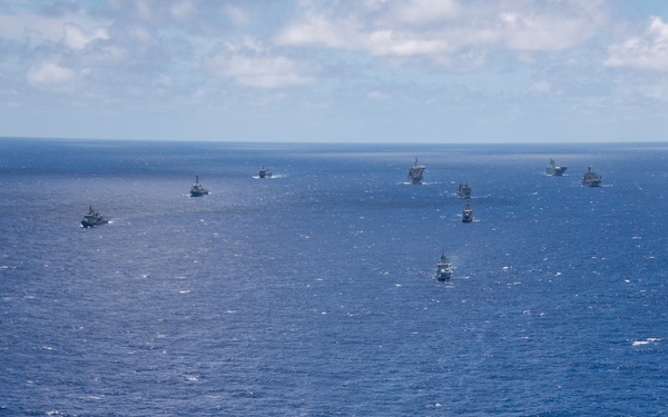 An aerial photograph shows a large naval formation of warships underway in open blue ocean under partly cloudy skies. The lead vessel in the foreground appears to be a Ticonderoga-class guided-missile cruiser, followed by numerous other warships extending toward the horizon in a column or multi-column formation. The scene is consistent with a multinational naval exercise or fleet transit operation.