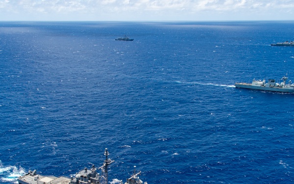 An aerial photograph shows a multinational naval formation underway in open blue ocean waters. The foreground features a large US Navy Ticonderoga-class guided-missile cruiser bearing hull number 57, accompanied by several other warships of varying sizes and apparent nationalities spread across the formation. The image appears to document a joint naval exercise or transit operation.
