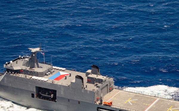 An aerial photograph of a military amphibious assault or landing platform dock (LPD) vessel underway at sea. The gray-hulled warship features a large flight deck at the stern with helicopter landing markings, and displays what appears to be a Philippine flag on its mast. The vessel is cutting through deep blue open ocean water at speed, generating a visible white wake.