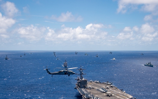 An aerial photograph shows a large nuclear-powered aircraft carrier underway in open blue ocean, surrounded by numerous escort and allied vessels spread across the horizon. A military helicopter, appearing to be an MH-60 Seahawk, flies alongside the carrier at close range. The scene appears to depict a multinational naval formation or fleet exercise, likely a RIMPAC or similar large-scale maritime exercise.