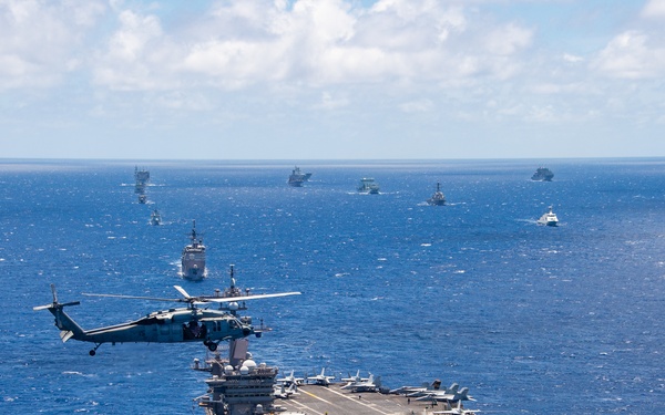 An aerial photograph shows a large nuclear-powered aircraft carrier underway in open blue ocean, surrounded by a substantial multinational naval fleet spread across the horizon. A military helicopter, consistent with an MH-60 Seahawk, is visible hovering or flying alongside the carrier's port side. Multiple fixed-wing aircraft are visible on the flight deck, suggesting this is a major naval exercise or fleet formation event.