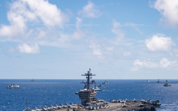 A large nuclear-powered aircraft carrier is underway in open blue ocean, surrounded by multiple escort vessels visible on the horizon. The carrier's flight deck is loaded with numerous fixed-wing aircraft arranged in organized rows. The formation suggests a carrier strike group conducting naval operations.