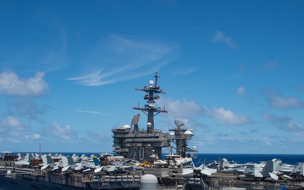 A large U.S. Navy aircraft carrier, visibly marked with the hull number '70' on its island superstructure, is underway in open blue ocean under partly cloudy skies. The flight deck is populated with numerous fixed-wing aircraft, and personnel are visible on the starboard sponson deck. The vessel appears to be in active operational status.