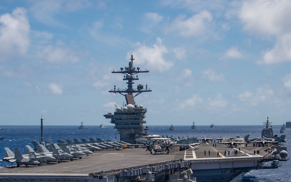 A large nuclear-powered aircraft carrier is underway in open blue ocean, surrounded by multiple escort and allied vessels visible on the horizon. The flight deck is populated with numerous fixed-wing aircraft arranged in rows, and personnel are visible on deck. The formation suggests a multinational naval exercise or transit operation.
