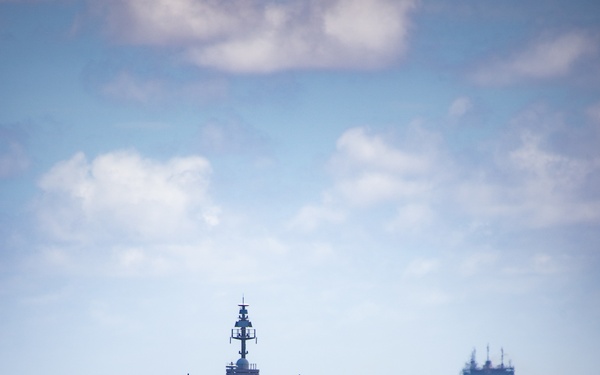 A formation of naval warships sailing in column formation across open blue ocean under a partly cloudy sky. The lead vessel is a large gray warship, identifiable by its distinctive superstructure, radar arrays, and dome-shaped sensor housings consistent with a Ticonderoga-class cruiser. At least three to four additional vessels are visible trailing behind in the distance.